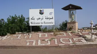 L'image montre un panneau de bienvenue à Fassouta, une localité. Le panneau est écrit en arabe et en anglais. À côté du panneau, il y a un abri qui semble abriter une statue, probablement de la Vierge Marie, indiquant une certaine importance religieuse à cet endroit. Le sol est décoré avec des mots formés de cailloux blancs, ajoutant une touche artisanale à l'ensemble. En arrière-plan, on aperçoit des arbres ou de la verdure, donnant une impression de nature environnante.