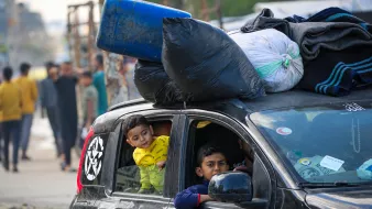 Sur cette image, on voit une voiture noire avec plusieurs objets attachés sur le toit, notamment des sacs et un bidon bleu. À l'intérieur du véhicule, il y a deux enfants. L'un des enfants, portant un vêtement jaune avec un imprimé, regarde par la fenêtre de la voiture. L'autre enfant, assis à l'avant, regarde également dehors. En arrière-plan, il y a plusieurs personnes qui semblent se déplacer, ainsi qu'un environnement urbain sans détails précis.