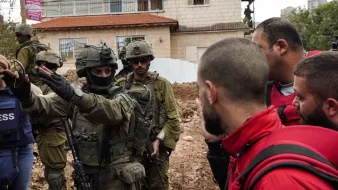 L'image montre des soldats en uniforme militaire, armés, entourés par un groupe de personnes. Certains sont en tenue d'urgence, tandis que d'autres semblent discuter avec les soldats. En arrière-plan, on voit des bâtiments et un terrain boueux. L'ambiance est tendue.