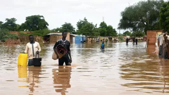 La imagen muestra una escena de inundación, donde el agua ha cubierto gran parte de un área, afectando a varias construcciones. Dos hombres caminan a través del agua, uno de ellos lleva un balde y el otro sostiene una manta enrollada. Al fondo, se pueden ver casas y árboles, todos rodeados de agua. El cielo está nublado, lo que sugiere una atmósfera sombría y húmeda. La inundación ha dejado a la comunidad en una situación difícil.