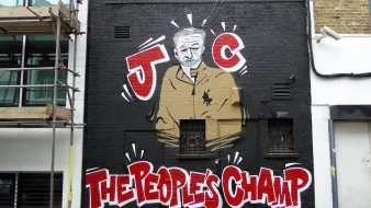 L'image montre un mur décoré d'un graffiti représentant une silhouette d'un homme en costume. Il est entouré de motifs graphiques et de couleurs vives. En bas, on peut lire les mots "THE PEOPLES CHAMP" écrits en lettres grandes et accrocheuses. Le graffiti est caractérisé par un style artistique urbain, typique des œuvres de street art. En arrière-plan, on aperçoit une construction, ajoutant un contexte urbain à l'œuvre.