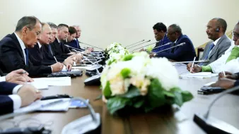 La imagen muestra una reunión formal en una sala con una mesa larga. Hay varias personas sentadas a ambos lados de la mesa, concentradas en la conversación. En el centro de la mesa, hay un arreglo floral decorativo. Las personas parecen estar intercambiando ideas, y algunos están tomando notas. El ambiente es serio y profesional.