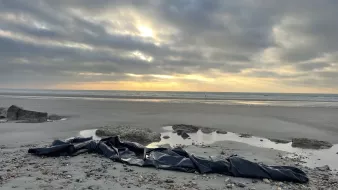 The image depicts a beach scene with a gray, cloudy sky creating a moody atmosphere. In the foreground, there is a large, black tarp or covering laid out on the sand, surrounded by smooth stones and scattered seashells. The beach appears to be wet, likely from recent tides, and there are gentle waves in the background. In the distance, a boat can be seen on the horizon, and rays of sunlight break through the clouds, creating a contrast with the darker tones of the sand and the tarp.