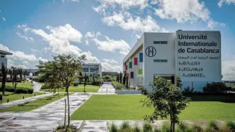 L'image montre l'Université Internationale de Casablanca. On peut voir des bâtiments modernes, avec des façades blanches et colorées. L'environnement est entouré de verdure, incluant des arbres et des espaces verts bien entretenus. Le ciel est bleu avec quelques nuages, et le sol semble humide, suggérant qu'il a peut-être récemment plu. L'ensemble dég dég dég dég dég dég dég dég dég dég dég dég dég dég dég dég dég dég dég dég dég dég dég dég dég dég deg d'une atmosphère accueillante et dynamique.