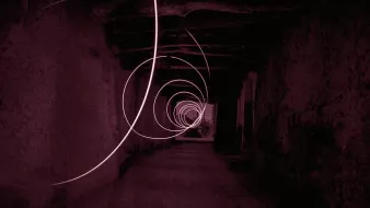 L'image présente un couloir sombre et étroit, avec des murs en pierre, qui semble s'étendre à l'infini. Des spirales lumineuses de couleur rose flottent dans l'air, créant une atmosphère mystérieuse et surréaliste. La lumière des spirales se détache sur le fond sombre, ajoutant une dynamique captivante à la composition. Le sol est en pierre, et la structure du plafond est visible, renforçant le sentiment d'un espace ancien ou abandonné.