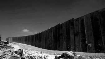 L'image montre un mur imposant s'étendant à l'horizon, dans un paysage désertique. Le mur est composé de grandes plaques de béton, parfois inégales. Au premier plan, il y a des débris et des éléments naturels, ce qui renforce l'impression d'un environnement aride et abandonné. Le ciel en arrière-plan semble dégagé, mais la scène est en noir et blanc, ce qui ajoute une atmosphère sombre et sérieuse à l'image.