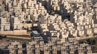 L'image présente un paysage urbain dense, constitué de nombreux bâtiments aux formes géométriques. Les structures sont principalement de couleur claire, avec des nuances de beige et de blanc. On peut observer des immeubles empilés les uns sur les autres, créant une apparence presque uniforme. Le terrain autour des bâtiments semble légèrement vallonné, avec des zones de végétation sporadique. Il y a également des éléments architecturaux distincts, tels qu'un toit qui se distingue parmi l'ensemble des constructions. L'ensemble donne une impression de modernité et d'urbanisation.