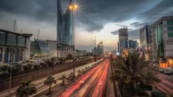 The image depicts a bustling urban scene, likely in a modern city. In the foreground, streaks of light from moving vehicles indicate heavy traffic on a multi-lane road, while palm trees line the sides, adding a touch of greenery. In the background, prominent skyscrapers with reflective glass facades dominate the skyline under a cloudy sky, creating a dramatic atmosphere. The scene captures a blend of modern architecture and vibrant city life, illuminated by streetlights and the fading light of sunset.