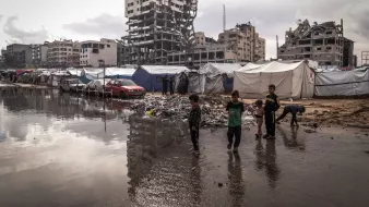 Des enfants jouent près de tentes dans un camp, avec un bâtiment en ruines en arrière-plan.