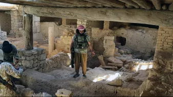 L'image montre un intérieur d'un bâtiment qui semble ancien et en ruine. On peut voir des murs en pierre et des débris éparpillés sur le sol. Un homme, habillé en vêtement militaire avec un bandeau, se tient au centre de la pièce, tandis que deux autres personnes, cachées en partie, l'observent. La lumière entre par des ouvertures, créant un contraste entre les ombres et les zones illuminées dans cet environnement désolé.