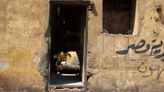 L'image montre une entrée partiellement ouverte d'un bâtiment avec un mur aux teintes jaunies et en mauvais état. À l'intérieur, on distingue un canapé doré, qui semble être un élément de mobilier abandonné. Le mur présente aussi des inscriptions en arabe, ajoutant une touche culturelle à la scène. L'ensemble dégage une atmosphère nostalgique, évoquant un espace autrefois habité et maintenant oublié.