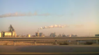 The image depicts an industrial landscape with several factories. In the foreground, there is a highway barrier, suggesting the viewer is looking from a moving vehicle. The factories emit smoke and steam into the atmosphere, creating white clouds against a clear blue sky. The overall scene conveys an industrial environment, typical of urban or semi-urban areas where manufacturing activities take place.