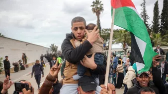 The image depicts a scene of a gathering or demonstration. A man is holding a child closely to him, showcasing a protective gesture. Around them, a crowd appears to be rallying, with some individuals raising their hands and a prominent Palestinian flag visible in the background. The atmosphere seems charged, indicating a moment of significance, possibly related to a political or social issue. The setting includes trees and a building in the background, against a cloudy sky.