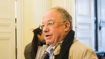 A man with glasses, light hair, and a tan coat stands indoors, speaking with someone behind him.