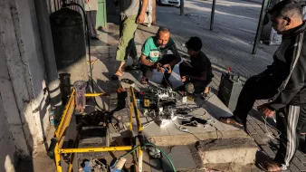 Nell'immagine si possono vedere alcune persone raccolte in un'area urbana. Tre uomini sono intenti a lavorare attorno a un dispositivo, probabilmente un generatore elettrico, che è posizionato su una base di cemento. Accanto al generatore ci sono vari cavi e dispositivi elettronici, suggerendo che stanno cercando di ricaricare batterie o alimentare apparecchi. Uno degli uomini è accovacciato mentre gli altri due stanno osservando o interagendo con i cavi. Sullo sfondo, si intravedono auto e strutture urbane tipiche. L'atmosfera sembra essere di lavoro e collaborazione nel contesto di un'area pubblica.