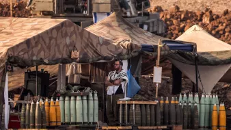 L'image montre un camp militaire en plein air. On y voit des tentes camouflées, et plusieurs obus de différentes tailles sont alignés sur des palettes. Un homme, possiblement en tenue religieuse, se tient debout près des tentes. En arrière-plan, on aperçoit un véhicule militaire. Le sol est poussiéreux, et le paysage semble aride, indiquant une zone de conflit ou d'entraînement militaire.