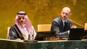 Two men at a conference table, one in traditional attire, discussing on a stage with microphones.