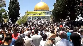 L'image montre une foule rassemblée devant un bâtiment orné d'un dôme doré. Les participants semblent être engagés dans une manifestation ou un rassemblement, avec des drapeaux et des pancartes visibles. Les arbres environnants ajoutent au décor, et l'atmosphère générale paraît dynamique, marquée par une forte présence de personnes.