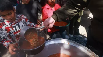 L'image montre des enfants qui se rassemblent autour d'une grande marmite. Un enfant, tenant une louche, verse une soupe dans un bol. D'autres enfants, attentifs, attendent leur tour pour recevoir leur repas. L'atmosphère semble conviviale, et l'environnement est extérieur, probablement dans une aire où des repas sont servis. Les enfants portent des vêtements variés, et certains semblent impatients et enthousiastes.