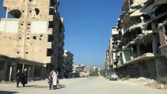 L'image montre une rue dévastée par des conflits, avec des bâtiments en ruines de part et d'autre. Les murs des bâtiments sont marqués par des impacts de balles et des débris sont éparpillés sur le sol. Au centre de la rue, quelques personnes marchent, tandis que des voitures abandonnées sont visibles. Le ciel est dégagé, ce qui contraste avec la destruction environnante.