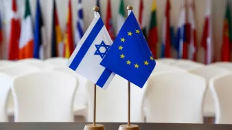 The image features two small flags standing on a table. One flag is the national flag of Israel, which has a blue Star of David and two horizontal blue stripes on a white background. The other flag represents the European Union, displaying a circle of twelve golden stars on a blue background. In the background, there are several other flags, likely from various countries, softly blurred out, suggesting a formal or diplomatic setting.