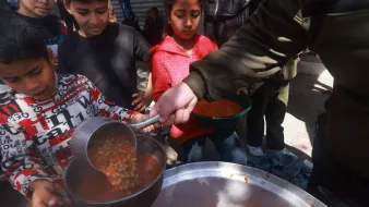 L'immagine mostra un gruppo di bambini in un ambiente all'aperto, mentre qualcuno sta servendo del cibo da una grande pentola. Un bambino, in particolare, sta utilizzando un mestolo per prendere una porzione di una zuppa o stufato, che sembra contenere legumi. Gli altri bambini attendono, con espressioni curiose e attente, alcuni con uno sguardo serio. L'atmosfera trasmette un senso di comunità e condivisione, evidenziando un momento significativo di aiuto e sostegno.