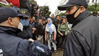 L'image montre une scène de tension entre des personnes en masse et des policiers. On peut apercevoir plusieurs individus, dont une femme portant un hijab et des vêtements clairs, qui semblent interagir avec les forces de l'ordre. Les policiers, en uniforme et portant des masques, sont en position d'encadrer la situation. L'arrière-plan montre une foule, indiquant un rassemblement ou une manifestation, et le temps semble pluvieux, ce qui ajoute à l'atmosphère de la scène.