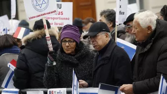 L'image montre une manifestation où un groupe de personnes tient des pancartes et des drapeaux. Les participants semblent se rassembler pour exprimer leurs opinions sur un sujet lié à Israël. Certains portent des signes indiquant des messages clairs, tandis que d'autres affichent des drapeaux français. L'atmosphère semble sérieuse et déterminée.