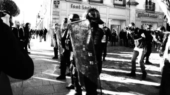 L'image montre une scène urbaine en noir et blanc, où des policiers en tenue anti-émeute, portant des casques et tenant des boucliers, se tiennent en position sur une rue. Ils sont face à une foule de personnes, dont certaines semblent prendre des photos. L'environnement est composé de bâtiments commerciaux, et l'ambiance semble tendue. Les ombres et la lumière créent un contraste saisissant dans la composition.