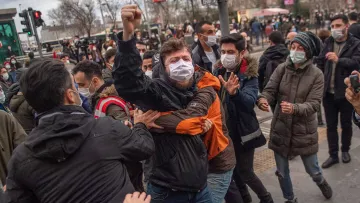 L'image montre une scène de tension où plusieurs personnes sont au centre d'une agitation. Un homme au premier plan lève le poing, exprimant possiblement une forte émotion. Les individus autour semblent en train de se disputer ou de se retenir, certains portant des masques. L'arrière-plan indique un environnement urbain avec des feux de circulation, ce qui suggère que la scène se déroule dans une ville. L'atmosphère paraît chargée, avec une dynamique de confrontation palpable.