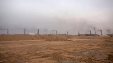 L'image montre un paysage industriel avec un sol désertique et nu. À l'arrière-plan, de nombreux cheminées émettent de la fumée dans une atmosphère brumeuse et chargée. Le ciel est grisâtre, ce qui donne une impression de pollution et de dégradation environnementale. Le terrain est principalement constitué de sable ou de terre, et l'ensemble dégage une atmosphère morose et industrialisée.