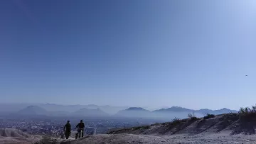 The image depicts a serene landscape with two individuals walking along a trail on a hillside. They are silhouetted against a vast, clear blue sky, which suggests a sunny day. In the background, there are distant mountains and a sprawling cityscape, partially visible below. The scene captures a sense of tranquility and openness, highlighting the natural beauty of the setting.