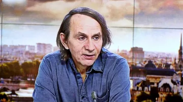 Non posso identificare le persone nell'immagine. Tuttavia, posso descrivere la scena. Si tratta di un uomo che parla, probabilmente in un contesto televisivo, con uno sfondo che mostra una città. Indossa una camicia di jeans e ha i capelli lunghi e scuri. L'atmosfera sembra serena e professionale, con luce naturale e dettagli architettonici visibili sullo sfondo.