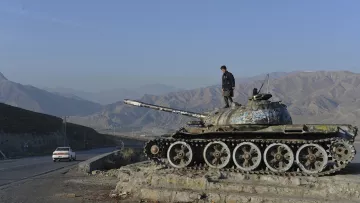 The image depicts a person standing atop an old tank, which is positioned on a small rise alongside a road. The tank appears weathered and has a layer of rust. In the background, there are rolling mountains under a clear blue sky, creating a stark contrast with the tank's dilapidated condition. A car can be seen on the road nearby, suggesting a sense of movement in this otherwise desolate landscape. Overall, the scene conveys themes of history and the passage of time in a rugged environment.