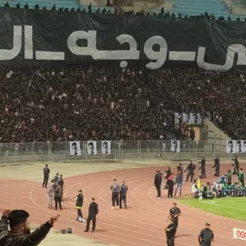 Stade plein de supporters en noir avec une grande banderole en arabe. Énergie et passion.