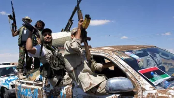 L'image montre des hommes armés debout sur un véhicule, probablement un pick-up, dans un environnement désertique ou semi-désertique. Ils affichent des signes de victoire ou de célébration, brandissant leurs armes au-dessus de leur tête. Le véhicule est couvert de boue et décoré de drapeaux ou d'insignes. Le ciel est dégagé, suggérant une journée ensoleillée. L'ensemble donne une impression de mouvement et de dynamisme, avec un contexte de conflit ou de célébration militaire.