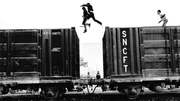 L'image montre un groupe de jeunes qui sautent d'un wagon de train à un autre. La scène est en noir et blanc, ce qui accentue l'action dynamique et l'intensité du moment. On peut observer deux jeunes sautant dans les airs, tandis que d'autres se trouvent sur les wagons ou à proximité. L'environnement semble urbaine, avec une atmosphère à la fois aventureuse et un peu risquée. Les détails des wagons et l'arrière-plan renforcent l'impression d'un cadre industriel.