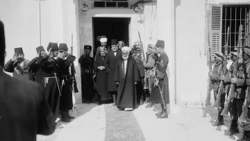 The image depicts a formal gathering outside a building, likely a historical site. A group of men dressed in traditional attire, including some wearing fezes and military uniforms, are seen saluting. In the center, an older man in a long robe and head covering is walking out of the entrance, suggesting he holds a position of significance or authority. The surrounding individuals, including soldiers and other men in formal dress, appear to be honoring or welcoming him. The scene conveys a sense of respect and formality typical of ceremonial occasions. The architecture hints at a historical and possibly Middle Eastern context.