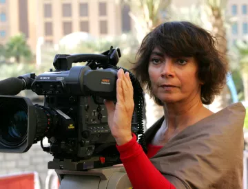 La imagen muestra a una mujer de cabello oscuro y rizado, que parece estar sosteniendo y operando una cámara de video profesional. Ella está mirando a la cámara con una expresión seria y concentrada. Viste una blusa color claro y tiene una capa o chal de un color más oscuro. Al fondo, se pueden ver edificios y un ambiente urbano. La escena sugiere que está en un evento o en el lugar de una grabación.