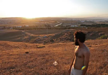 L'image montre un homme debout sur une colline, regardant vers un paysage vallonné au coucher du soleil. Il a les cheveux et la barbe bruns, et porte un t-shirt. Le terrain autour de lui est sec et couvert d'herbes hautes. En arrière-plan, on peut voir un petit village et des champs qui s'étendent à perte de vue, créant un panorama paisible et naturel. La lumière du soleil met en valeur les couleurs dorées de la scène.