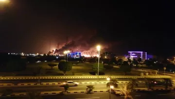 L'image montre une scène nocturne où l'on peut voir des nuages de fumée s'élevant dans le ciel, suggérant qu'il y a un incendie ou une explosion quelque part à l'horizon. Les lumières des bâtiments environnants sont visibles, créant un contraste avec l'obscurité de la nuit. La route devant est éclairée, avec quelques voitures qui passent. Des arbres et des espaces verts se trouvent également au premier plan, ajoutant une certaine profondeur à la scène.