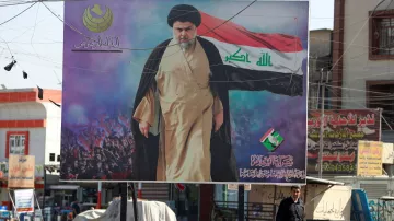 در این تصویر یک بیلبورد بزرگ نشان داده شده است که یک مرد با لباس روحانی و مسئولیت‌های سیاسی را در حال حرکت به سمت دوربین به تصویر می‌کشد. او دست خود را بلند کرده و یک پرچم در پس‌زمینه وجود دارد که نشان‌دهنده نمادها و پیام‌های خاصی است. در پس‌زمینه، جمعیتی از مردم در حال تماشا و حمایت هستند. فضای عمومی تصویر نشان‌دهنده یک منطقه شهری است که شاید تحت تأثیر مسائل اجتماعی یا سیاسی باشد. 