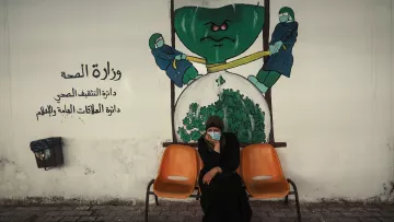 L'image montre une femme assise sur un banc orange, dans une ambiance urbaine. Elle porte un vêtement traditionnel noir et a un masque sur le visage. Derrière elle, on peut voir une fresque murale représentant deux personnages en tenue de protection, qui semblent tirer sur une grande horloge en forme de verre à sablier contenant du matériel de santé, symbolisant probablement une thématique liée à la santé publique. Le texte en arabe indique "Ministère de la Santé". L'ensemble de la scène évoque des questions de santé et peut-être de vigilance face à des défis sanitaires.