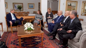 The image depicts a formal meeting taking place in a well-lit, elegantly furnished room. A group of professionals is seated around a coffee table, engaged in discussion. On one side, a person in a suit appears to be leading the conversation, while others listen attentively. The room features tasteful decor, including paintings on the walls and a floral arrangement on the table, creating a calm and sophisticated atmosphere. The group consists of both men and women, indicating a diverse representation.