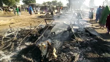 L'image montre une scène de destruction, où des débris brûlés et des objets en métal tordu sont dispersés sur le sol. De la fumée s'élève de restes calcinés, suggérant qu'un incendie a eu lieu récemment. À l'arrière-plan, on aperçoit des personnes en train de se déplacer, sur un terrain qui semble être une communauté ou un marché. L'atmosphère est sombre et le paysage est marqué par les dégâts causés par le feu.