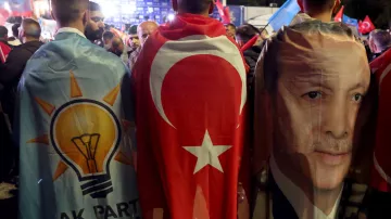 L'image montre un groupe de personnes réunies lors d'un événement politique, enveloppées dans des drapeaux et des bannières. Deux d'entre elles portent des capes : l'une présente le symbole du parti AKP (Parti de la justice et du développement), tandis que l'autre arbore le drapeau turc et une image du président Erdoğan. On peut voir une ambiance de rassemblement avec des drapeaux turcs en arrière-plan, suggérant une atmosphère de soutien et de célébration.