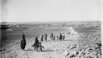 The image depicts a group of people walking along a rocky path in a barren landscape. They appear to be headed toward a distant town or village, which is visible in the background. The group consists of several adults, some of whom are carrying burdens on their heads, and a child walking alongside them. The terrain is dry and rugged, with scattered rocks and sparse vegetation. The sky is clear, and the scene conveys a sense of movement and migration through a rural setting.