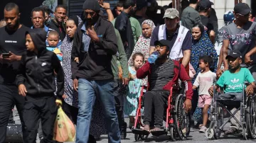 L'image montre une scène de rue animée, remplie de personnes de différents âges. On peut voir des adultes marchant, certains portant des enfants. Un homme en fauteuil roulant est également présent, buvant de l'eau. La diversité des vêtements et des expressions faciales souligne une ambiance de vie quotidienne dans un environnement urbain. En arrière-plan, on aperçoit des bâtiments et une foule d'autres personnes.