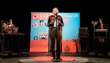 L'image montre une scène de spectacle, probablement un cabaret. Au centre, un artiste souriant est sur scène avec un micro, habillé de manière théâtrale. En arrière-plan, un grand panneau coloré affiche le mot "CABARET" et "COLONISATION", suggérant un thème spécifique. De chaque côté, on aperçoit des musiciens ou des techniciens avec des instruments ou des équipements, contribuant à l'atmosphère festive et engageante. Le sol est orné d'un tapis rouge, ajoutant une touche de mise en scène au spectacle.