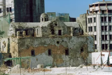 L'image montre des ruines d'un bâtiment ancien, probablement une église, dont la structure est en pierre et partiellement en décomposition. La façade est endommagée et recouverte de végétation, tandis qu'en arrière-plan, on peut voir des immeubles modernes en construction ou en rénovation. Le contraste entre l'ancien et le moderne donne une atmosphère de dégradation et de changement architectural. La zone semble être en chantier, avec des barrières et des échafaudages visibles.
